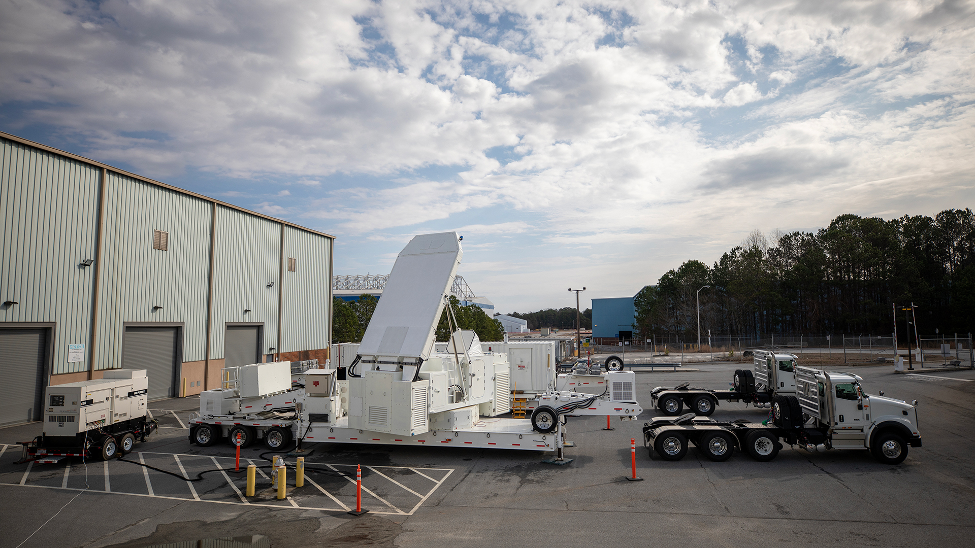 Large white device on the bed of a tractor trailer next to a large warehouse.