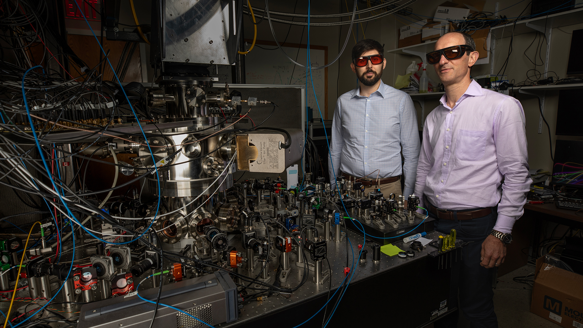 Two male researchers standing in a darkened room next to a large electronic device with many parts and wires.