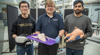 Three young male researchers standing side by side, left one holding VR glasses, center one holding raw whole chicken, right one holding rubber chicken 